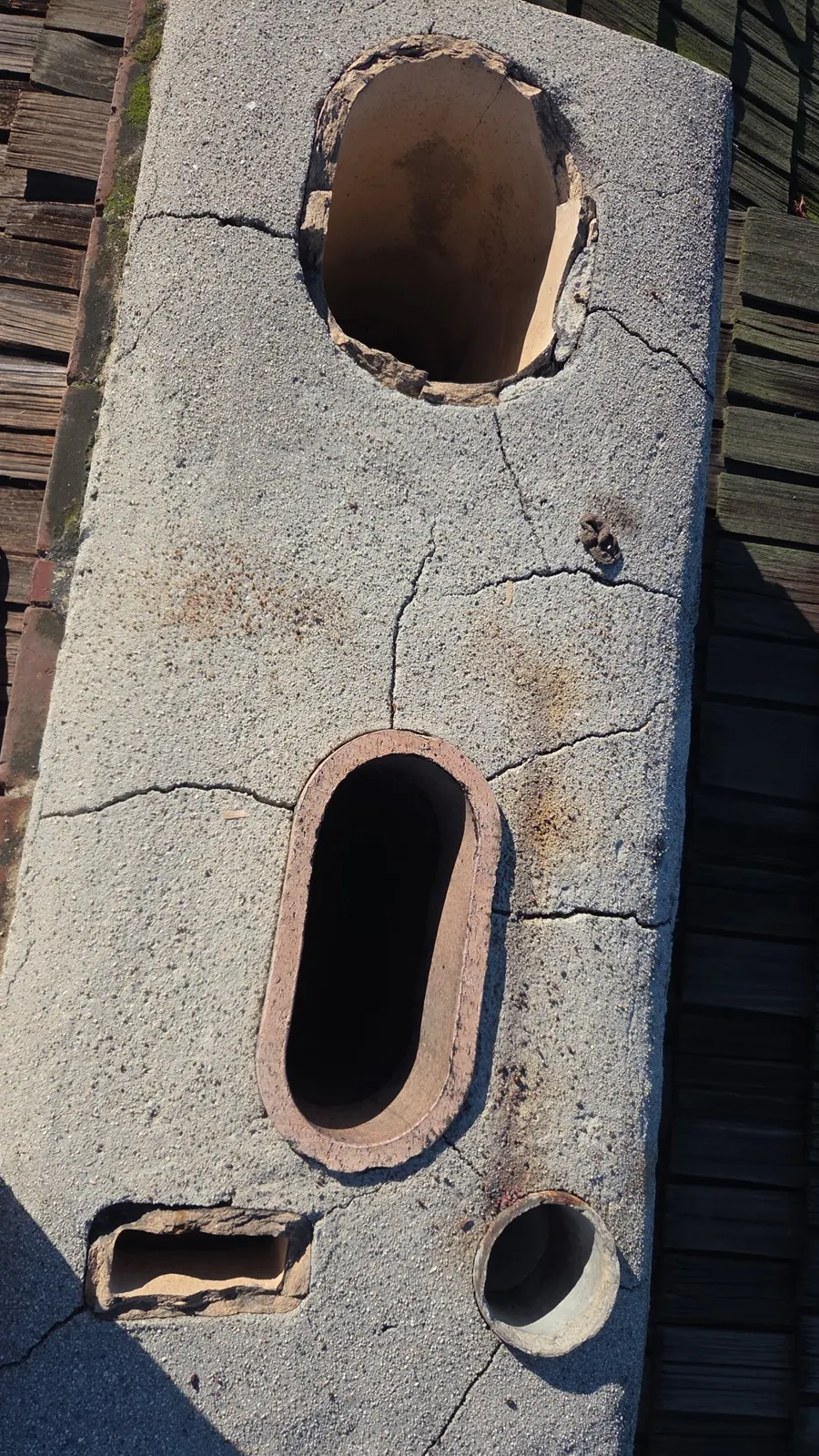 Cracked concrete chimney crown with chipped clay flue tiles and multiple flue openings, needing chimney repair, seen from above on a wood shake roof