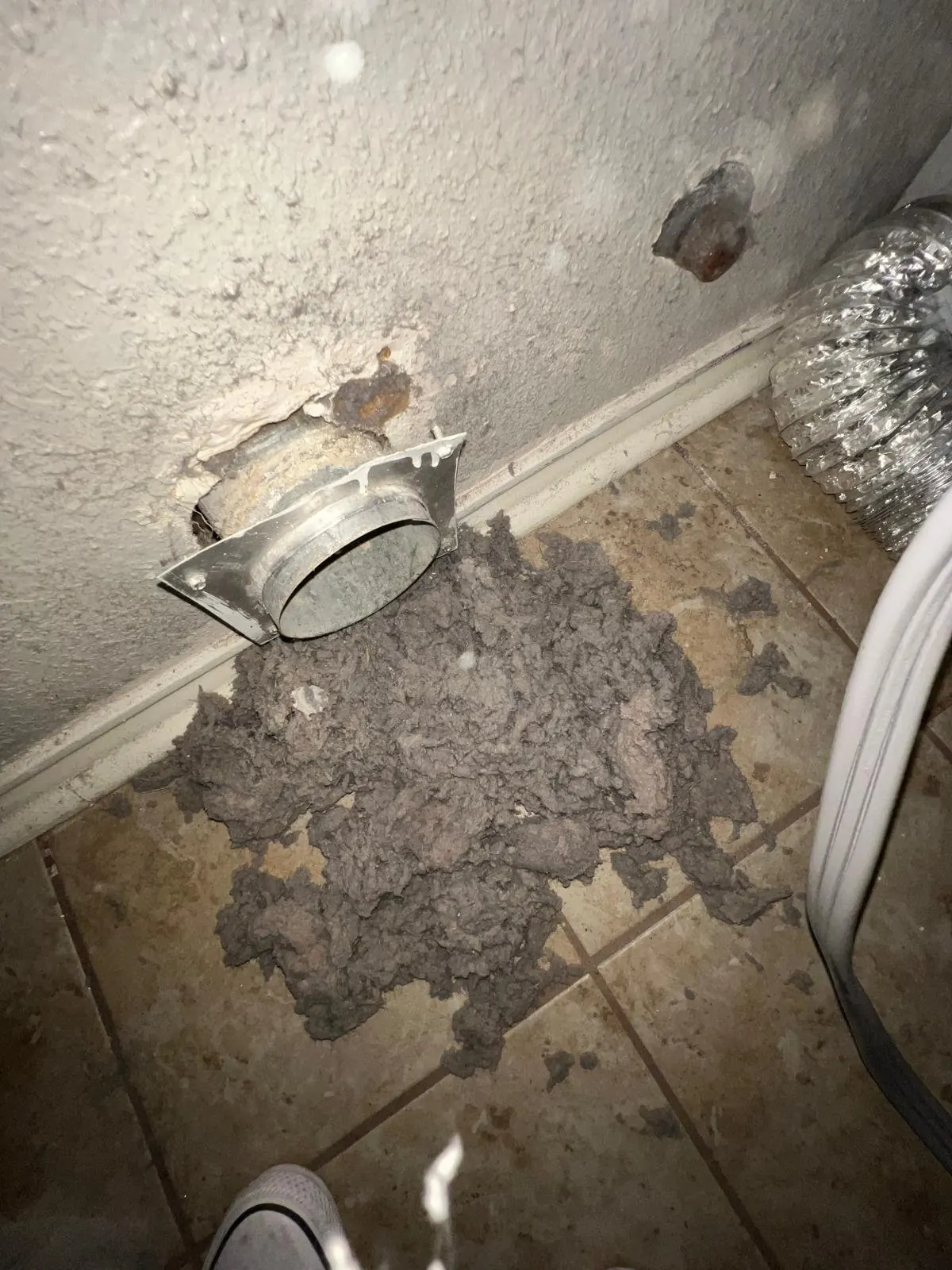 Large pile of lint removed from a clogged dryer vent, lying on the tile floor below the wall vent opening during a dryer vent cleaning